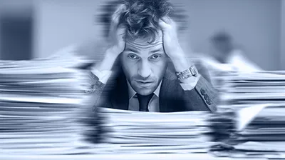 Dirigeant dépassé, la tête entre les mains, entouré de piles de dossiers, illustrant la surcharge de travail du dirigeant de petite structure.